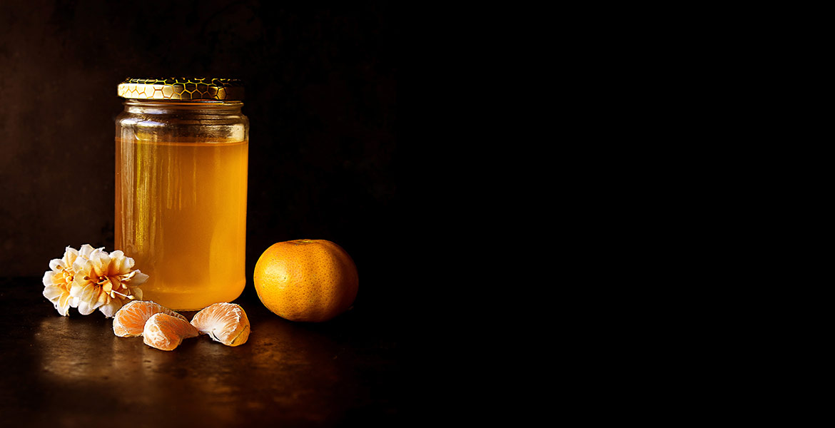 A jar full of honey next to an orange, orange slices, and a flower.